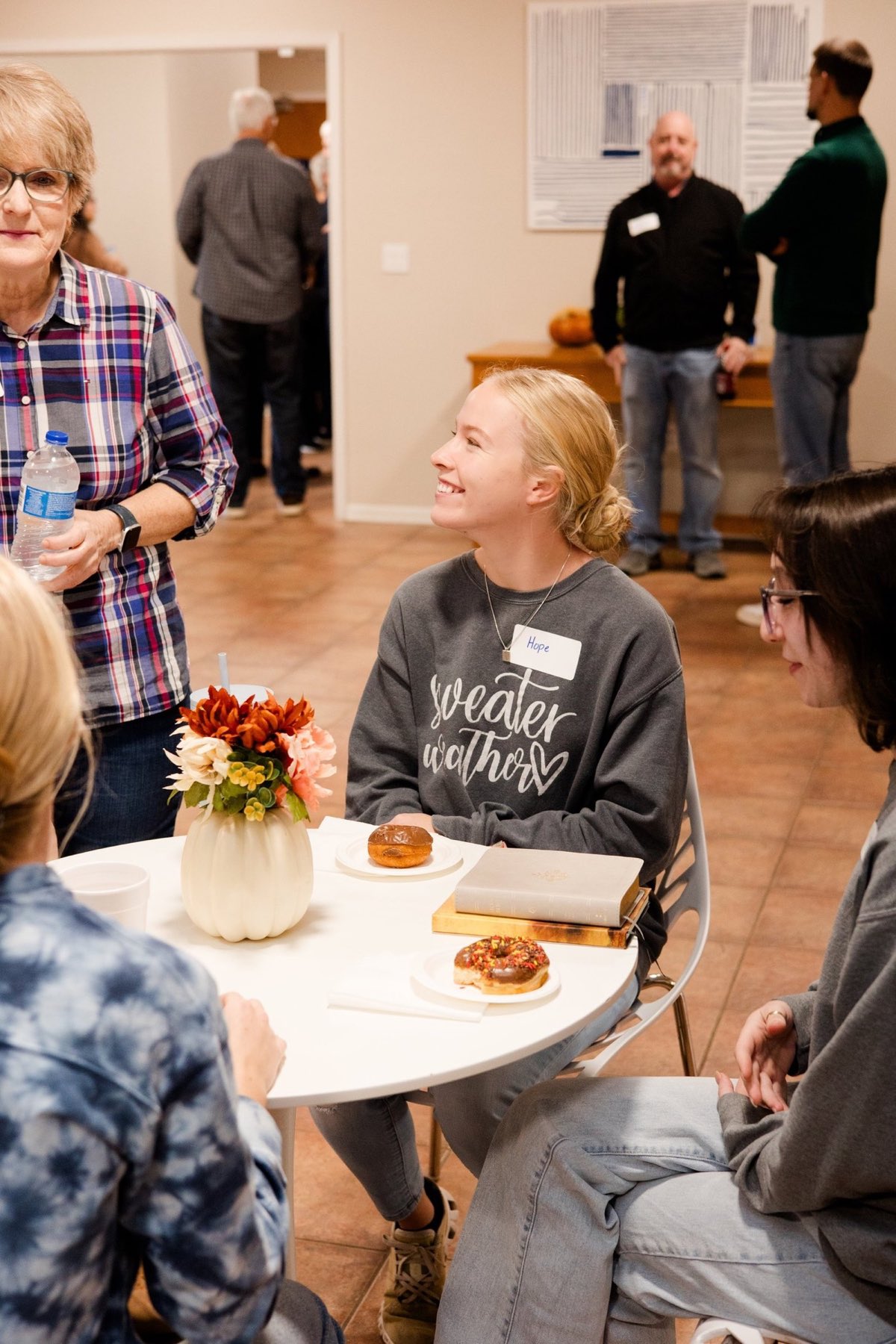 A guest wearing a name tag smiles at a table with donuts and a Bible during Sunday morning coffee and breakfast.