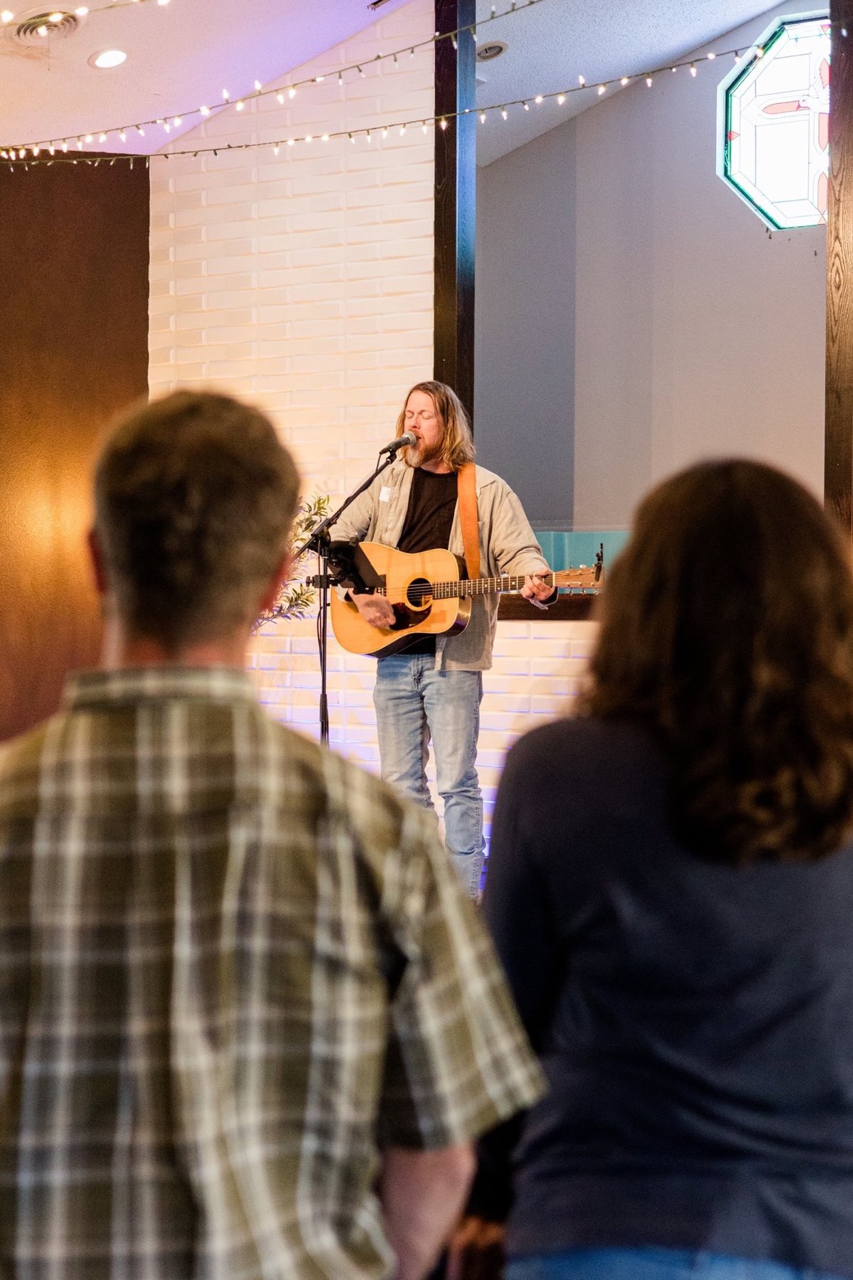 A worship leader plays guitar and sings on stage while two people stand watching in the foreground