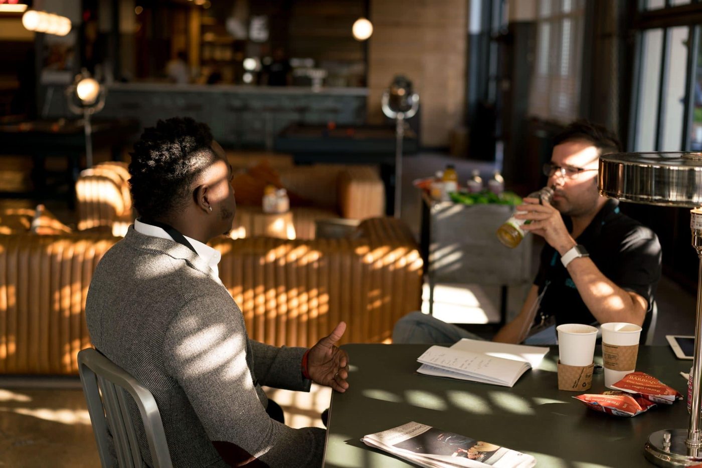 Two men talking together at a table in a cafe, with coffee cups and notebooks in front of them